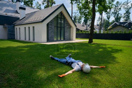 Téléchargez les photos : Constructeur en casque dur et gants en caoutchouc couché sur la pelouse dans la cour en face de la maison privée nouvellement construite - en image libre de droit