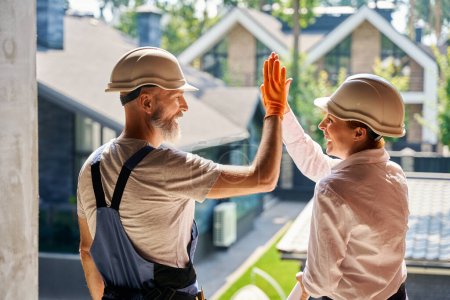 Téléchargez les photos : Inspecteur de bâtiment joyeux donnant haute-cinq à constructeur heureux à l'intérieur de la maison résidentielle inachevée - en image libre de droit