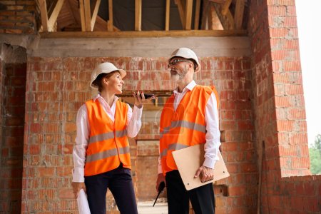 Photo for Joyful building inspector talking on smartphone while standing next to contractor inside unfinished residential house - Royalty Free Image