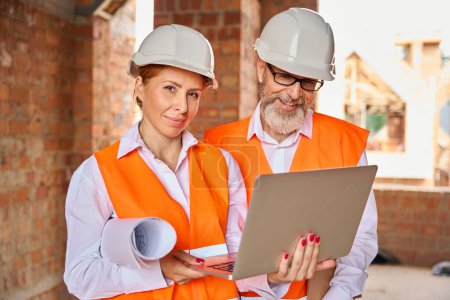 Photo for Female engineer showing something on laptop to her joyful colleague in uncompleted residential building - Royalty Free Image