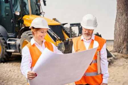 Photo for Smiling female architect and foreman looking at architectural drawings while standing in front of backhoe loader - Royalty Free Image