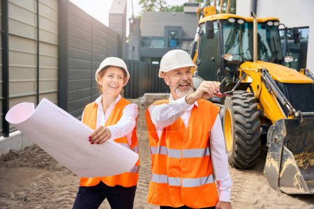 Photo for Cheerful contractor demonstrating something in distance to smiling construction supervisor while standing near backhoe loader - Royalty Free Image