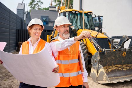 Photo for Foreman showing something in distance to female building inspector while standing near backhoe loader - Royalty Free Image