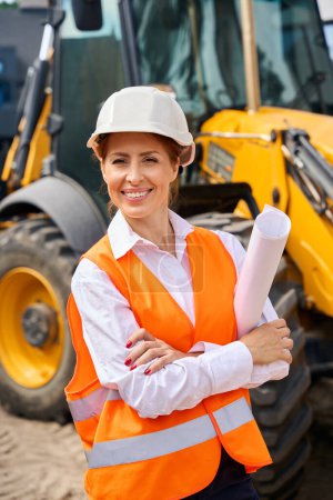 Photo for Waist-up portrait of cheerful engineer with rolled-up blueprints under arm standing near backhoe loader - Royalty Free Image