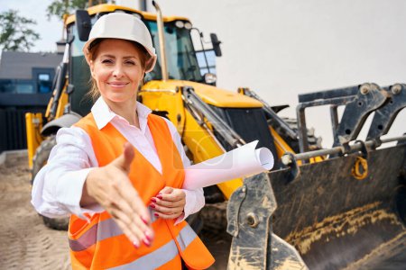 Photo for Waist-up portrait of smiling construction manager extending hand for handshake with somebody while standing near backhoe loader - Royalty Free Image