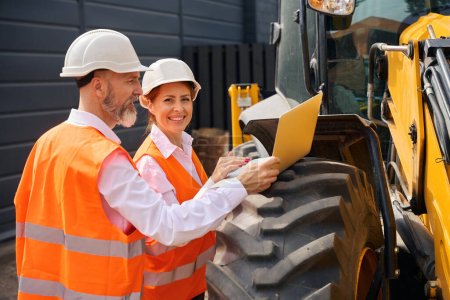 Photo for Smiling female engineer and her focused colleague using computer placed on backhoe loader wheel outdoors - Royalty Free Image