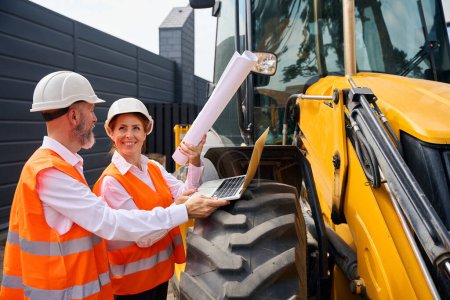 Photo for Smiling female engineer with architectural drawings in hand pointing up at something while talking with foreman near backhoe loader wheel - Royalty Free Image