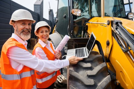 Photo for Joyous foreman with laptop and female engineer with blueprints standing outdoors near backhoe loader wheel - Royalty Free Image