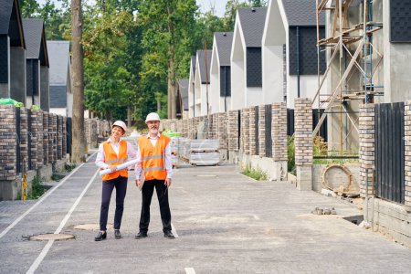 Photo for Full-length portrait of smiling engineer with blueprints and pleased foreman with walkie-talkie standing on paved street among unfinished houses - Royalty Free Image