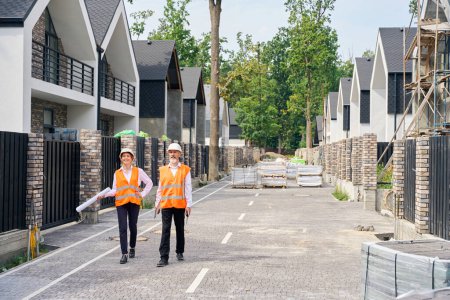 Photo for Full-size portrait of pleased female engineer and her colleague walking along paved street with unfinished private residential houses - Royalty Free Image