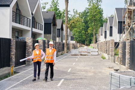 Photo for Full-length portrait of engineer and female architect walking along paved street among unfinished private residential houses - Royalty Free Image