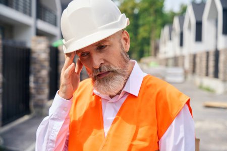 Photo for Portrait of exhausted construction superintendent in hard hat touching his temple while standing on street among residential houses - Royalty Free Image