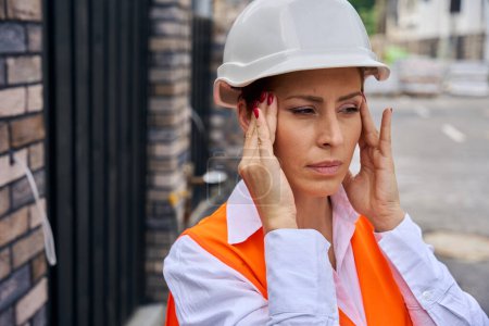Téléchargez les photos : Inspecteur de bâtiment femme épuisée en casque dur toucher ses tempes avec les mains tout en se tenant à l'extérieur pendant l'attaque de migraine - en image libre de droit