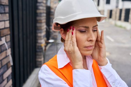 Photo for Portrait of tired construction supervisor in hard hat touching her temples with hands during migraine attack on street - Royalty Free Image