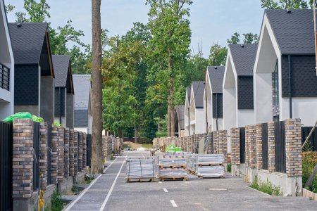 Téléchargez les photos : Rangées de nouvelles maisons privées modernes le long de la rue pavée avec des matériaux de construction sur des palettes en bois - en image libre de droit