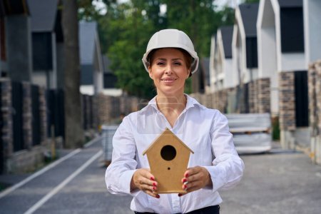 Photo for Waist-up portrait of pleased exterior designer with wooden nesting box in her hands standing between rows of houses - Royalty Free Image
