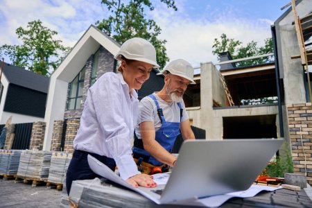 Photo for Contented female engineer and housebuilder looking at architectural drawings on building plot - Royalty Free Image