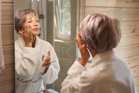 Foto de Agradable dama en un albornoz se aplica crema en la cara, se mira en un espejo grande - Imagen libre de derechos