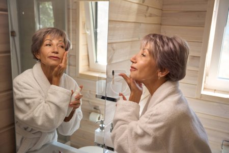 Photo for Elderly woman applies cream to her face in front of a mirror, the bathroom is bright and clean - Royalty Free Image