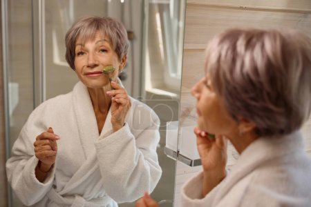 Téléchargez les photos : Femme âgée en peignoir utilise un masseur facial jade, la salle de bain est propre et confortable - en image libre de droit