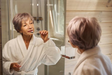 Photo for Elderly woman brushes her teeth with a toothbrush in front of a mirror, the bathroom is bright and clean - Royalty Free Image