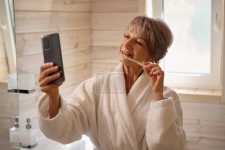 Téléchargez les photos : Dame en peignoir prend un selfie avec une brosse à dents, la femme a un téléphone portable moderne - en image libre de droit