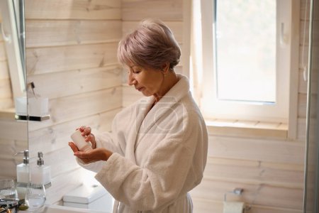 Téléchargez les photos : Souriante dame âgée prend des vitamines du matin dans la salle de bain, elle a beaucoup de produits de soins de la peau - en image libre de droit