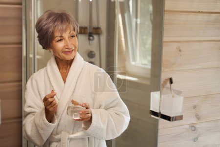 Téléchargez les photos : Belle dame âgée prend des vitamines du matin dans la salle de bain, elle tient un verre d'eau - en image libre de droit