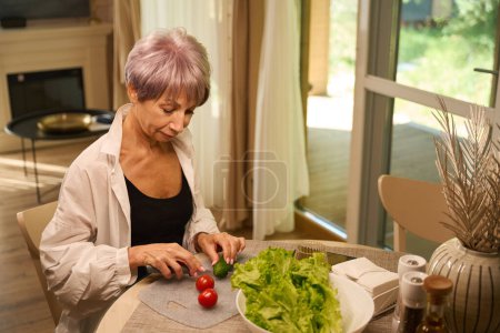 Photo for Elderly lady is preparing vegetable salad from fresh vegetables in her kitchen, there is a decorative vase on the table - Royalty Free Image