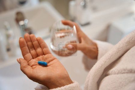 Foto de La mujer sostiene una píldora analgésica y un vaso de agua en la mano, ella está en el baño - Imagen libre de derechos