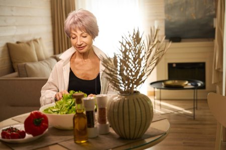 Foto de Señora jubilada está preparando ensalada de verduras frescas en su cocina, hay un jarrón decorativo en la mesa - Imagen libre de derechos