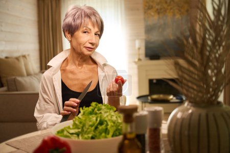 Photo for Pensioner prepares a vegetable salad from fresh vegetables in her kitchen, there is a decorative vase on the table - Royalty Free Image