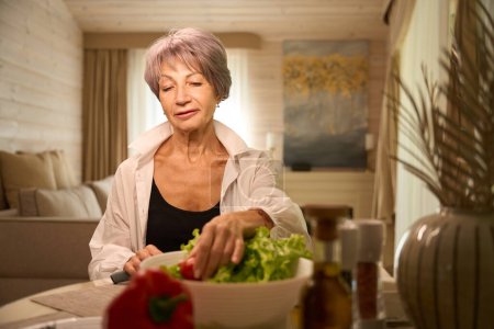 Elderly pensioner is preparing a vegetable salad in her kitchen, there is a decorative vase on the table