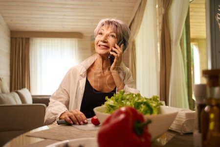 Téléchargez les photos : Belle femme âgée communique sur un téléphone portable dans sa cuisine, elle prépare une salade de légumes - en image libre de droit