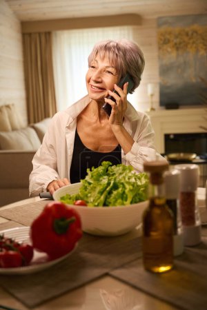 Foto de Niza anciana se comunica en un teléfono móvil en su cocina, ella prepara una ensalada de verduras - Imagen libre de derechos
