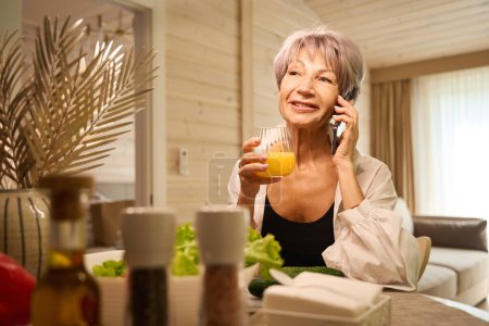Téléchargez les photos : Femme âgée communique sur un téléphone portable dans sa cuisine, elle a un verre de jus dans ses mains - en image libre de droit