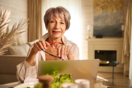 Foto de La anciana con una camisa blanca trabaja remotamente en su cocina, usa un portátil - Imagen libre de derechos