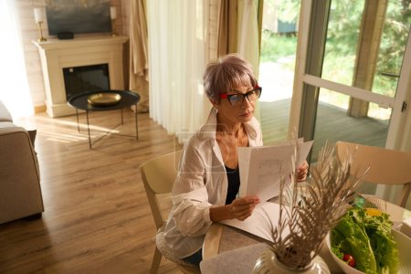 Photo for Female works remotely in her kitchen with work papers, she uses a laptop - Royalty Free Image