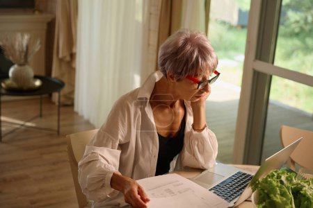 Téléchargez les photos : Femme dans une chemise blanche à la maison dans la cuisine avec un ordinateur portable et des papiers de travail, légumes frais sur la table - en image libre de droit