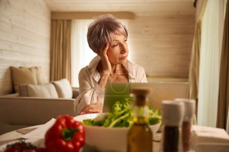 Foto de Mujer cansada trabaja remotamente en su cocina, utiliza un ordenador portátil - Imagen libre de derechos
