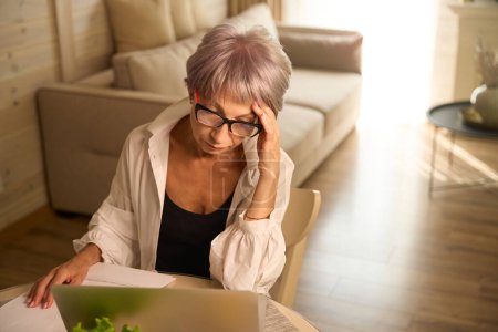 Téléchargez les photos : Femme avec des lunettes travaille à distance dans sa cuisine, elle utilise un ordinateur portable - en image libre de droit