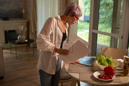 Téléchargez les photos : Femme âgée à la maison dans la cuisine avec un ordinateur portable et des papiers de travail, légumes frais sur la table - en image libre de droit