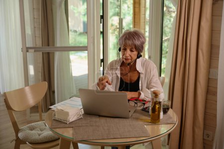 Foto de Anciana se comunica en línea en su cocina, ella utiliza un auricular especial - Imagen libre de derechos