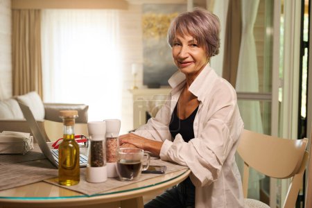 Téléchargez les photos : Femme assise à la table de la cuisine avec un ordinateur portable, huile et épices sur la table - en image libre de droit