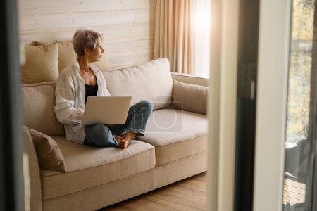 Foto de Mujer bonita en ropa casual se sienta con un ordenador portátil en un sofá acogedor, en una sala de diseño ecológico - Imagen libre de derechos