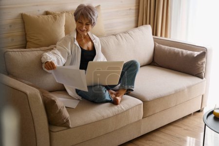 Photo for Smiling woman with a short haircut sits with a laptop on a cozy sofa in an eco-design room - Royalty Free Image