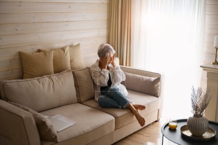 Ältere Frau leidet unter Kopfschmerzen, sie sitzt zu Hause auf einem gemütlichen Sofa