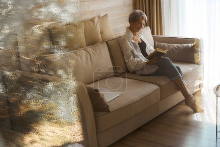 Téléchargez les photos : Dame âgée souriante se détendre avec un livre sur un canapé confortable, à côté d'un ordinateur et des documents de travail - en image libre de droit
