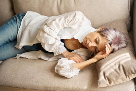 Téléchargez les photos : Femme âgée s'allonge pour se reposer sur un canapé confortable, elle souffre d'un mal de tête - en image libre de droit