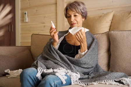 Photo for Elderly female is suffering from a cold, she is sitting on the sofa with a nasal spray and a napkin - Royalty Free Image